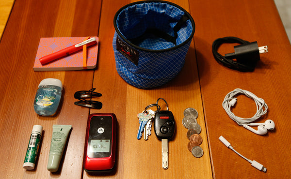 A Small Travel Tray surrounded by various accoutrements, a charge cable, earbuds, a dongle, a bit of change, a pair of keys, a notepad and pen, lotion, chapstick, hair-clips and a flip phone.